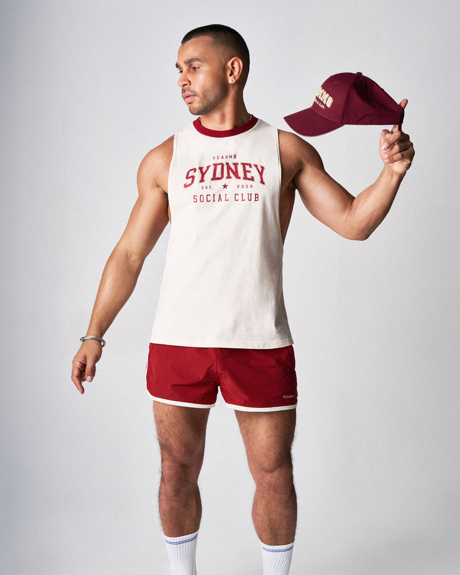 Man wearing a 'Sydney Social Club' tank top and red shorts, holding a cap.